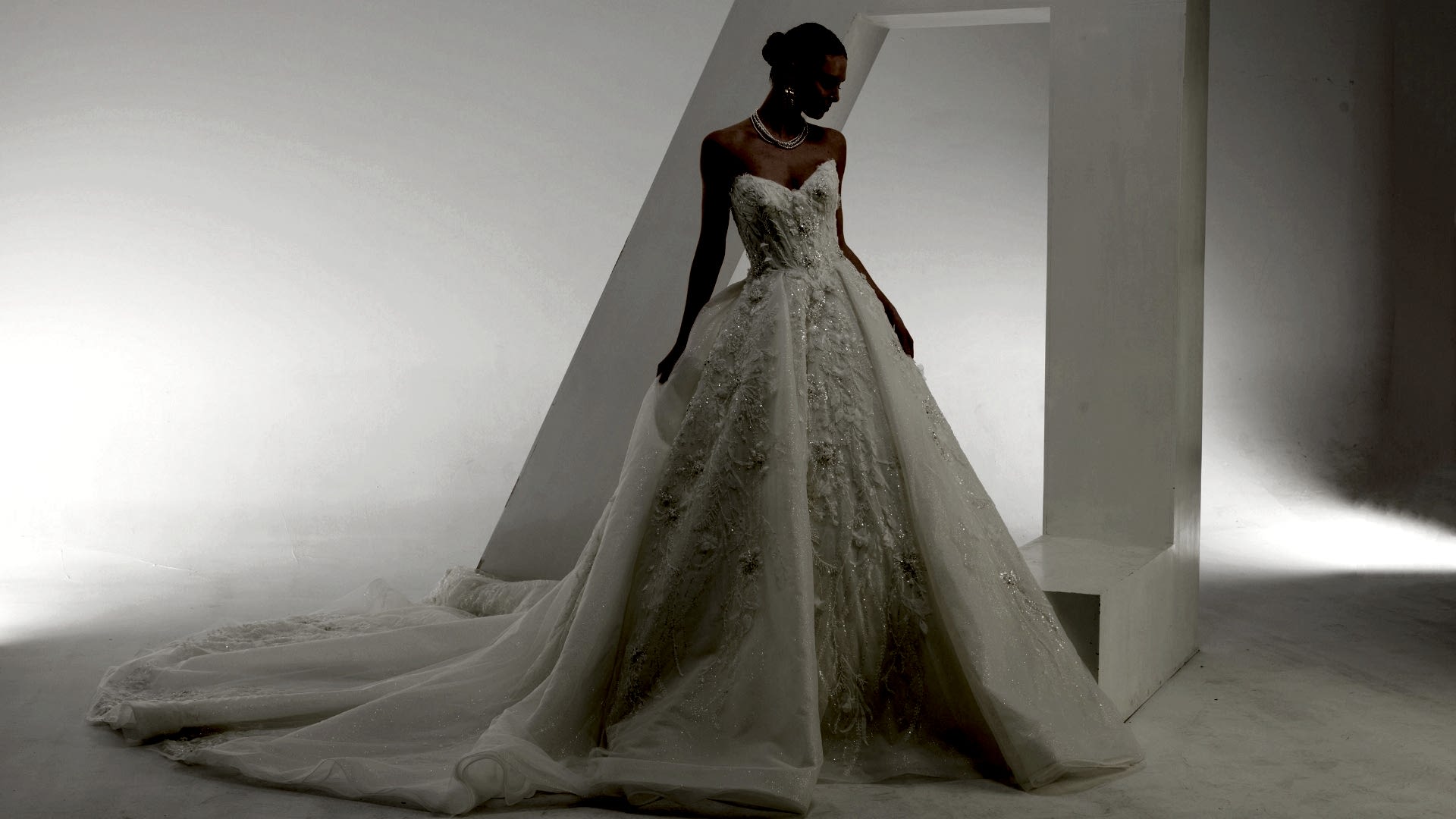 Elegant bridal gown with a voluminous ballgown skirt, intricate 3D lace appliqué, and a structured corset bodice, worn by a smiling bride in a modern white setting.