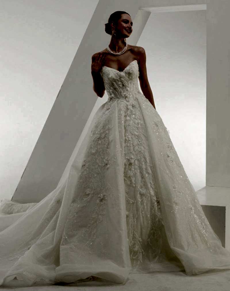Elegant bridal gown with a voluminous ballgown skirt, intricate 3D lace appliqué, and a structured corset bodice, worn by a smiling bride in a modern white setting.